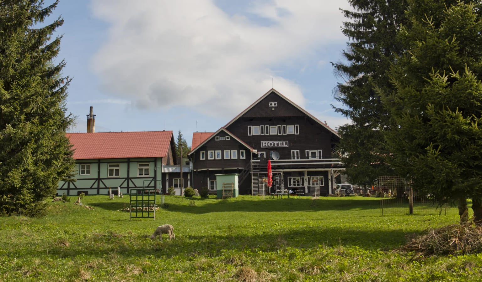 Hotel Červena Liška – tmavá dřevěná budova s nápisem HOTEL a sousední stavení s červenou střechou na zelené louce mezi smrky